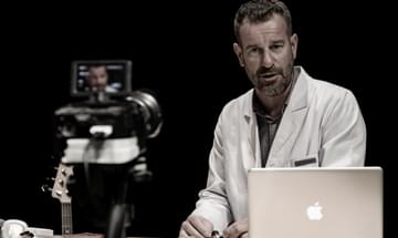 A man in a doctor's coat is sat at a desk with a laptop in front of him, looking into a video camera