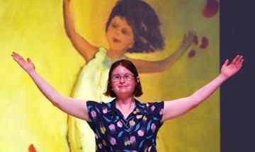 A woman with Down Syndrome stands with her arms open wide in front of a painting of a child with her arms in the same position.