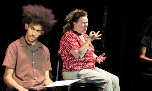 A man with an afro sits looking off to the side, whilst a woman next to him speak sign language