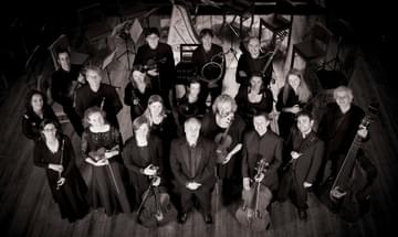 A black and white group photo of the orchestra ensemble all dressed up for concert, standing and looking up at the camera while holding their instruments in their hands