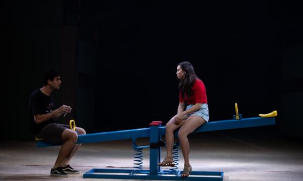A young man and woman sit on stage on a blue seesaw, deep in conversation