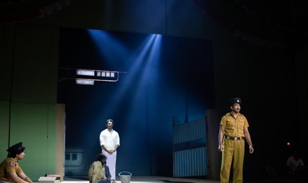 A prison guard stands looking out at the audience from the front of the stage whilst a dishevelled prisoner sits on the floor far behind him.