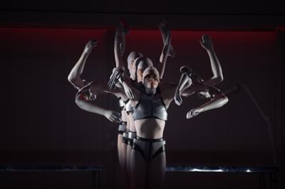 A line of dancers dressed as robots hold artificial limbs in a spider formation