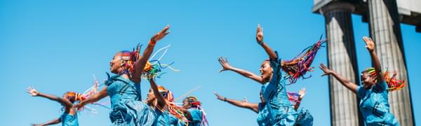 A group of women in bright blue African wax print dresses and colourful braids dance on top of Calton Hill