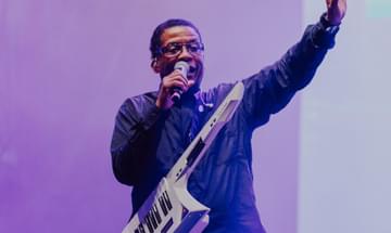 A man stands with a microphone in one hand and his other arm raised with a keytar around his neck in front of a purple background.