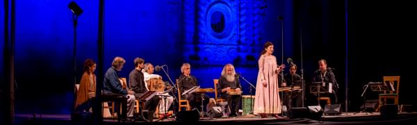 An ensemble of musicians playing traditional Turkish instruments sit in a semi-circle around a woman who is standing and singing, in front of a castle lit up in blue.