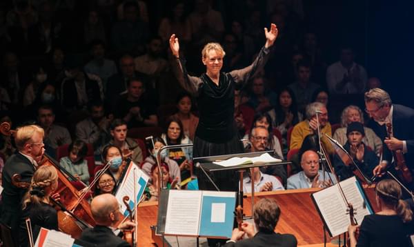 A blonde woman stands conducting an orchestra with her arms raised and a smile on her face