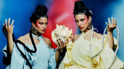 Two woman in matching puffy dresses, one blue and one yellow, sit holding a conch shell in one hand and their braids in the other.