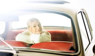 A woman sits inside a car with her head resting over her arms and looking out from the back of the car to the camera