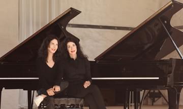 The sisters sit together on a piano stool with two grand pianos behind them, both dressed in black with red lipstick on.