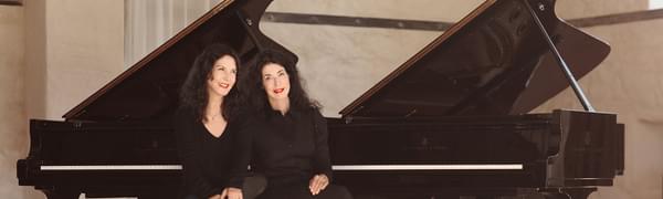 The sisters sit together on a piano stool with two grand pianos behind them, both dressed in black with red lipstick on.