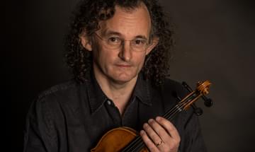 A man with shoulder-length curly hair, wearing a black shirt and wire-rimmed glasses, sits holding a violin, looking up at the camera.