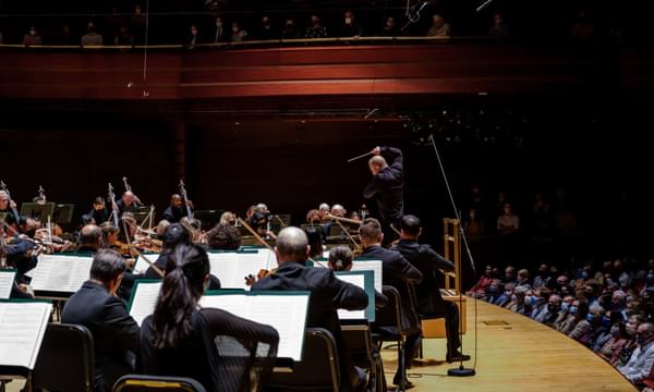 A conductor is standing with his right arm in the air in front of a full orchestra on a stage, with an audience looking on.