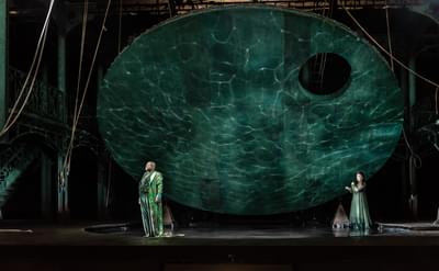 A man and a woman stand on stage under a large suspended disk with watery projections behind them.