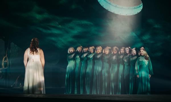 A group of water nymphs stand in a pool on stage with a woman in a white dress stood apart looking at them