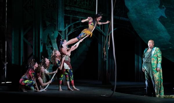 A tower of aerialists are all balanced along a rope, with a man in a jacquard metallic jacket stood singing on the right-hand side