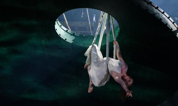 A woman in a white dress is suspended from above through a hole in the roof, leaning back and extending her arm.