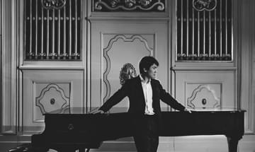 A greyscale photo of Seong Jin Cho dressed in a black suit leaning against a grand piano with his arms stretched out to both side.