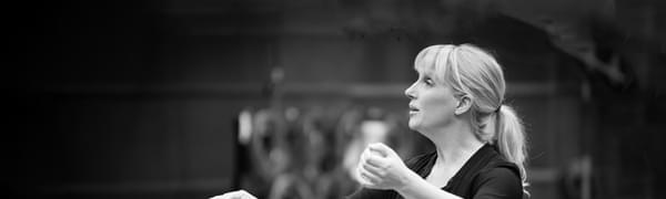 A black and white photo of a woman dressed in black with her hands raised and ready to conduct. She looks away from the camera to the musicians outside of the picture