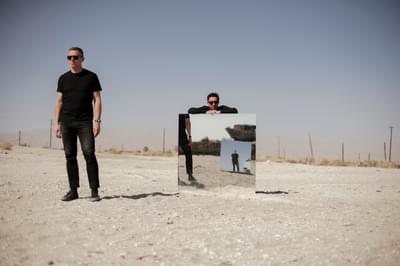 Two men stand in a desert, one of them leaning on a large mirror. You can see a reflection of them in another mirror.