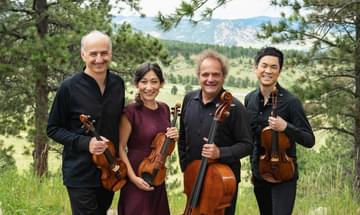 A string quartet stand in a grassy field against a mountainous backdrop, smiling towards the camera and holding their instruments.