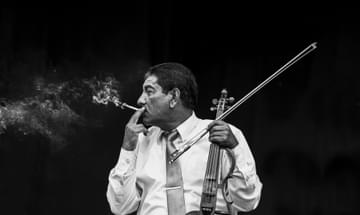 A greyscale photo of a man sat with his head turned to the side, smoking a cigarette with plumes of smoke emerging from it with an electric violin resting on his knee.