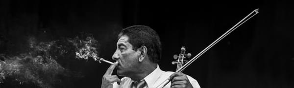 A greyscale photo of a man sat with his head turned to the side, smoking a cigarette with plumes of smoke emerging from it with an electric violin resting on his knee.
