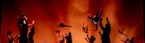 Lots of gymnasts are performing jumps and tumbles in silhouette against a red, smoky background.