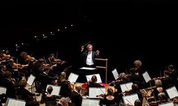 A conductor with his arms aloft in front of a sea of string instrument musicians