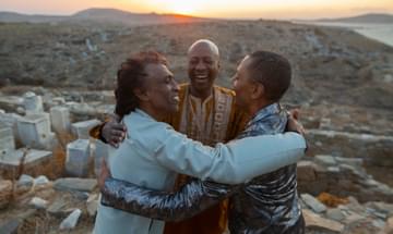 Three men stand in a desert landscape embracing and smiling at each other