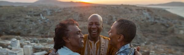 Three men stand in a desert landscape embracing and smiling at each other