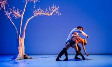 A man and a woman are dancing in a blue room with a cardboard tree.