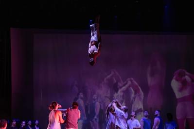 An acrobat flies high in the air, upside down with his body straight and arms by his side, with others waiting below to catch him and a large screen behind showing a different perspective on the staging.