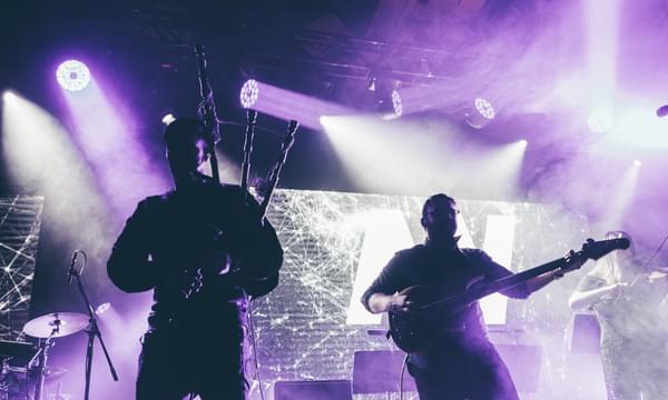 A piper, electric guitarist and fiddler perform on a hazy, strobe-lit stage, with microphones and drumkits visible around them.
