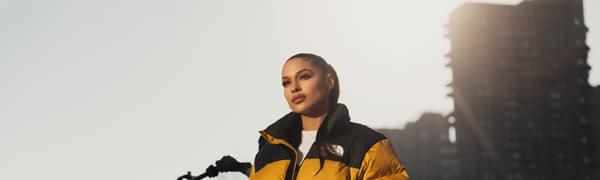 A woman stands in early-morning sunlight looking down into the camera, with her hair slicked back into a low ponytail and her hands in the pockets of her yellow puffa jacket. A bicycle wheel and tower block are visible behind here.