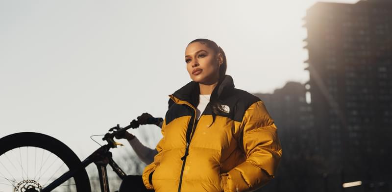 A woman stands in early-morning sunlight looking down into the camera, with her hair slicked back into a low ponytail and her hands in the pockets of her yellow puffa jacket. A bicycle wheel and tower block are visible behind here.