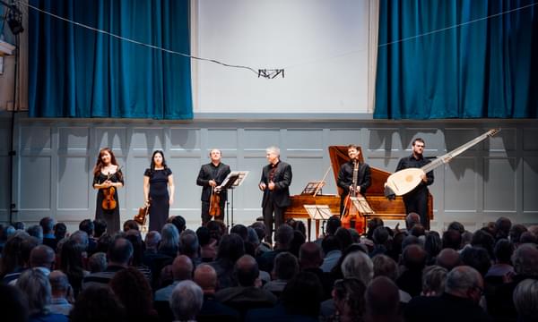 Six musicians in black stand on the stage with their instruments after a concert. The walls and curtains behind them are blue and in front of them a full concert hall of people is applauding.