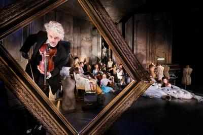 A man with curly white hair stands in a large picture frame, holding a violin. Behind him many people sit on a stage, surrounded by mannequins