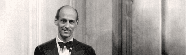 A black-and-white photograph of a man wearing a bow tie and black tuxedo, standing in a wood-panelled room andsmiling at the camera