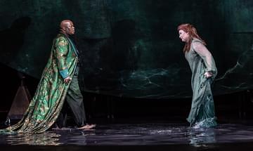 A man and woman, both dressed in elaborate robes, sing to each other on a dark, watery stage, facing each other with intent expressions.
