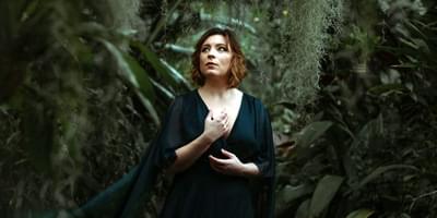 A woman in a flowing green dress stands in a dark forest with moss trailing around her. She is looking off to the side.