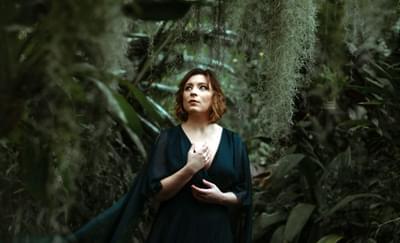 A woman in a flowing green dress stands in a dark forest with moss trailing around her. She is looking off to the side.