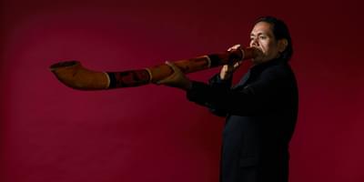 A man dressed in a black suit stands against a maroon background, playing an ornately decorated didgeridoo
