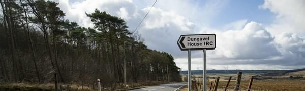 An image of a road on a sunny day, with a forest on one side and fields on the other with a road sign pointing to 'Dungavel House IRC'