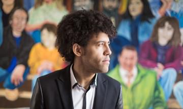 A man with light brown skin and short curly hair sits in the middle of the image wearing a suit and shirt. He looks to the side and behind him is a colourful, blurred out mural of lots of sitting people.