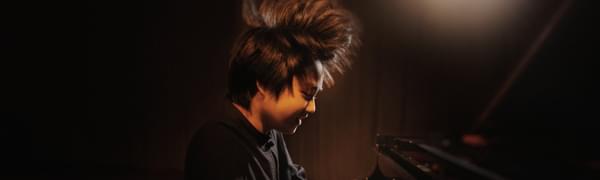 Mao Fujita plays a grand piano furiously, so much so that his hair stands on end. He is bathed in a spot of warm orange light