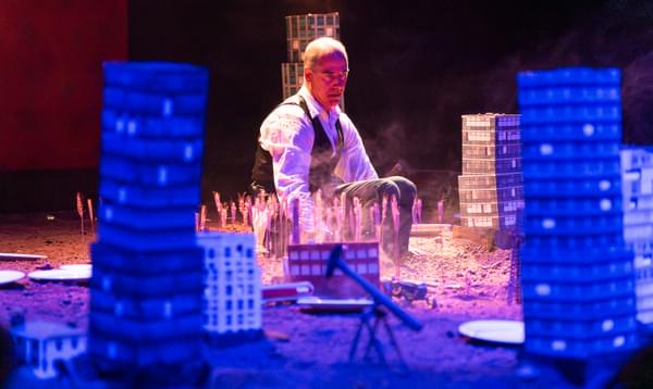 An actor wearing a white shirt and waistcoat sits immersed in dirt. Around him are model city buildings, trucks and oil rigs., tr