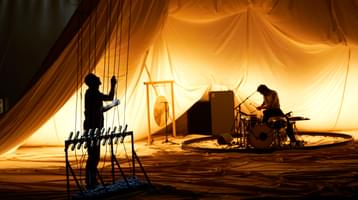 In a billowing orange tent, two musicians are cast in shadow. One sits at a drum set and the other plays an instrument that has strings suspended from the ceiling