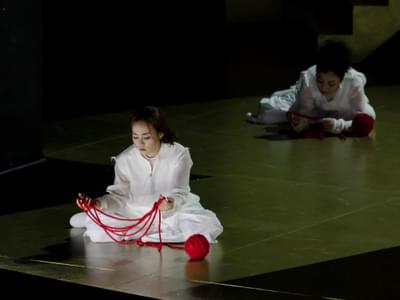 Two women in white shifts sit on the floor playing with strands of red yarn. One is illuminated by white light while the other sits in darkness.