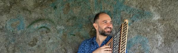 A man stands against a wall holding a zither and smiling